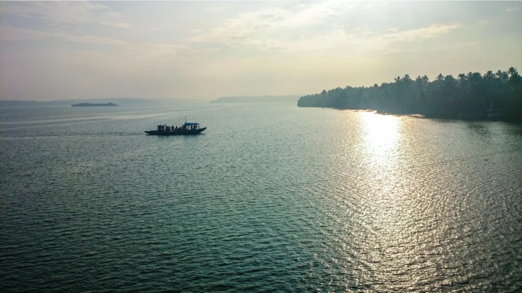 Ashtamudi Lake in Kollam with a boat cruising across the calm waters during sunset, surrounded by lush coconut trees.