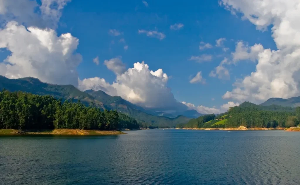 Mattupetty Dam in Munnar, Kerala, surrounded by tranquil blue waters, lush forests, rolling hills, and dramatic clouds against a clear sky.