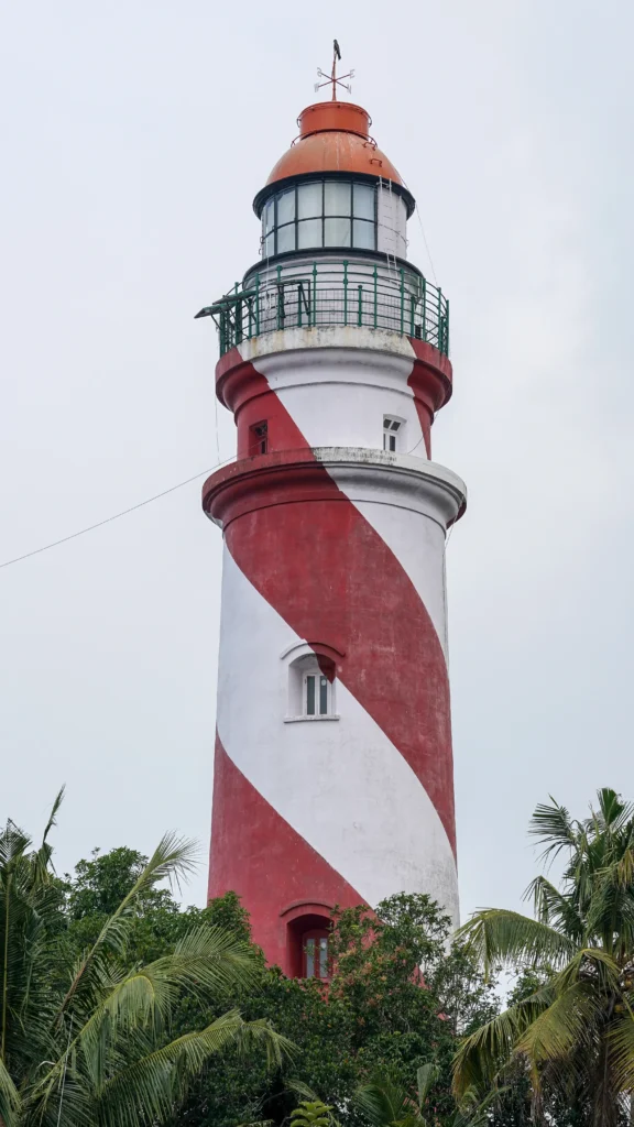 Thangassery Lighthouse in Kollam, a tall red-and-white striped lighthouse surrounded by coconut trees under a cloudy sky.