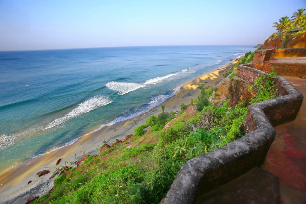 Aerial view of Varkala Beach in Kerala, showing a wide sandy shoreline, blue Arabian Sea, coastal cliffs, and lush palm trees.